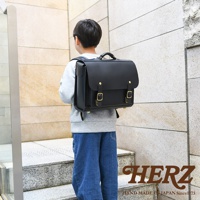 男孩背著黑色皮革橫式書包，書包上有金屬扣環，下方有HERZ品牌字樣。