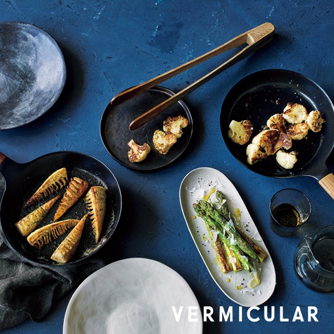 日本製黑色琺瑯鑄鐵平底深鍋，鍋內有烤過的根莖類蔬菜。旁邊有烤過的花椰菜、蘆筍，以及木質夾子和餐盤。