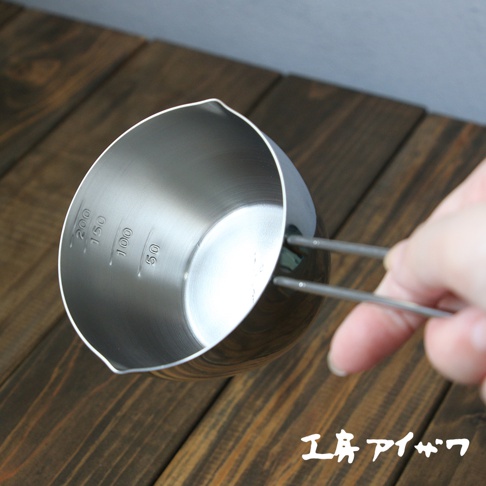 不鏽鋼長柄量杯，內側標有 50、100、150、200ml 的刻度，杯緣有傾倒口，由手持細長的不鏽鋼手柄。