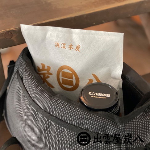一個灰色織布紋理的相機包袋內，裝著一包印有出雲屋炭八字樣與品牌標誌的白色方形調濕木炭包，旁邊則放置了一顆黑色機身的佳能單眼相機鏡頭，鏡頭蓋上印有 Canon 與 Ultrasonic 字樣。
