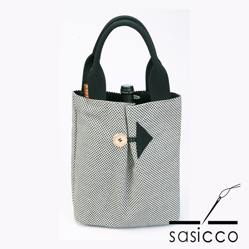 一個直立的黑色和白色刺子織托特包，配有黑色提把。包的正面有一個木製鈕扣和一個黑色箭頭形狀的裝飾。包內有一個酒瓶。