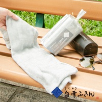 一條淺灰色紗布長巾，質地輕薄，帶有織紋，一角由手拿起。旁邊是一個透明塑膠袋包裝的同款長巾，袋上印有日文標籤。背景為木製長凳與綠色植物，右側有一副太陽眼鏡。