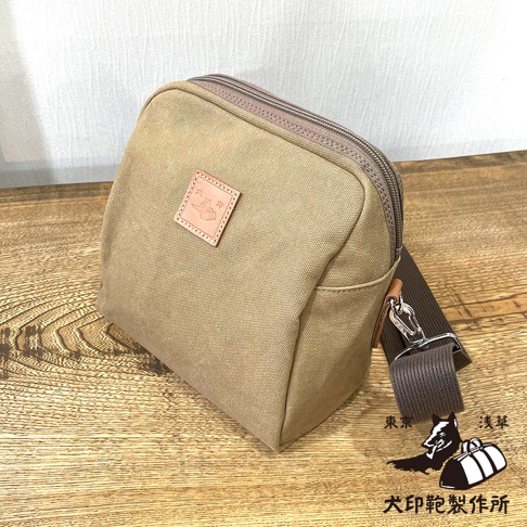卡其色石洗帆布製成的大容量散步包，包身方正，頂部有拉鍊開合，前方有一個方形的皮革標籤，上面壓印著圖案和文字。包包側面有深棕色的帆布背帶，掛鉤為銀色金屬。背景是淺色紋理牆面和木紋桌面。右下角有兩個黑色的圖案，一個是站立的狗，另一個是兩個行李袋，下方有日文文字。