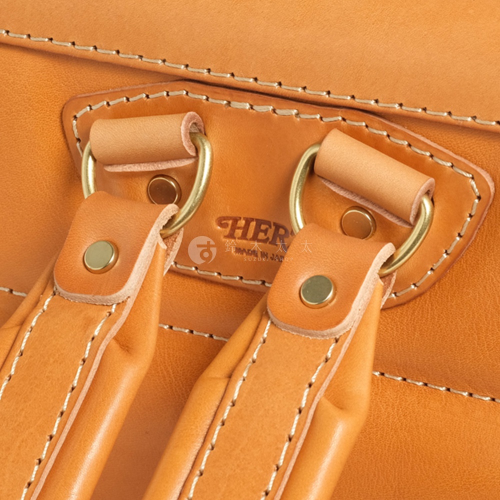 職人手作皮革橫式書包的細節，包含皮質的縫線、金屬環扣和皮革提把，部分可見品牌標誌。