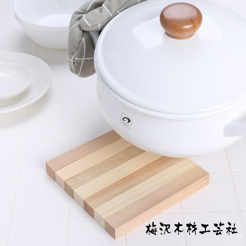 由多塊淺色木條拼接而成的方形隔熱墊，擺放著白色鍋具，鍋蓋上有圓形木製把手。