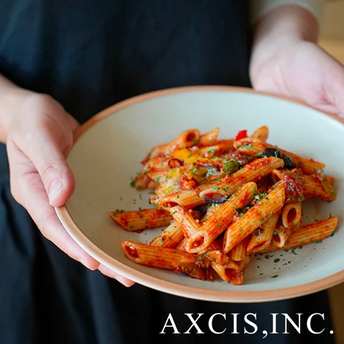 一盤裝有番茄醬汁、蔬菜和香草的筆管麵，盛裝在一個淺色的圓形陶瓷義大利麵盤中，盤緣有著淡粉色裝飾。