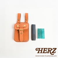 一個橘棕色皮革製成的經典腰袋，帶有黃銅扣環和兩個掛環，旁邊放著一支深灰色電子煙和一盒綠色薄荷口味加熱式煙草。