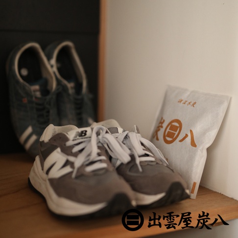 木質層架上放置著一包印有出雲屋炭八標誌的白色調濕木炭包，前方擺放一雙灰白色 New Balance 運動鞋，背景另有一雙藍色運動鞋，整體呈現居家生活的收納場景。