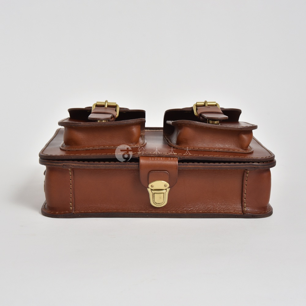 職人手作咖啡色皮革雙口袋兩用單肩包，正面有兩個翻蓋口袋，以金色金屬扣環固定，中間有一金色旋轉鎖扣。