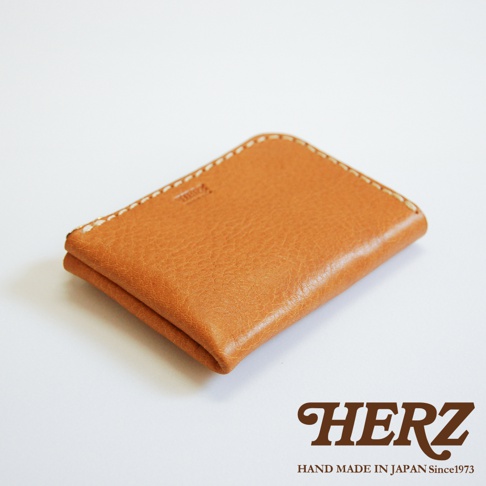 這是一款日本HERZ職人手工製作的皮革卡片收納夾，採用柔軟的Stare牛皮革製成，呈現自然的暖棕色澤。外觀設計簡約俐落，配有精緻的米白色縫線細節與品牌標誌。其L型收納開口設計方便拿取物品，整體輕便耐用，下方印有HERZ品牌名稱與成立年份。