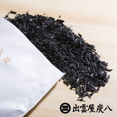一張俯視照片，顯示出雲屋炭八小型室內調濕木炭，深黑色的針葉木炭顆粒從白色不織布包裝袋中傾倒而出，散落在淺木色平滑表面上，展示天然木炭的細碎質感與品牌標誌。