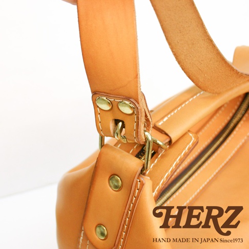 職人手作皮革厚實單肩波士頓包，展現天然皮革的溫潤色澤與紮實紋理，金屬扣件與車縫線細節清晰可見，品牌Logo「HERZ」與「HAND MADE IN JAPAN Since1973」字樣點綴其中。