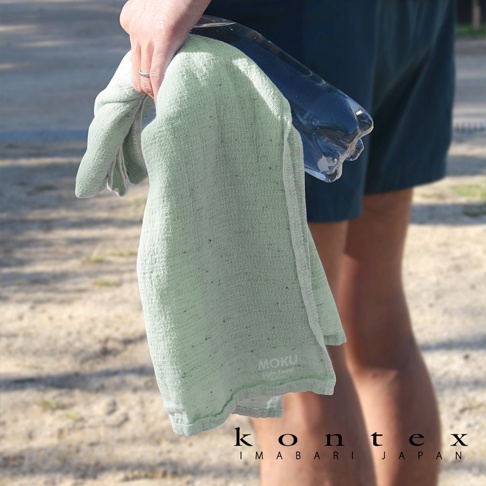 MOKU超速乾輕薄吸水浴巾，淺綠色，輕柔材質，帶有細緻紋理，一角印有MOKU品牌標誌，被一隻手握住，旁邊有一瓶透明水瓶。