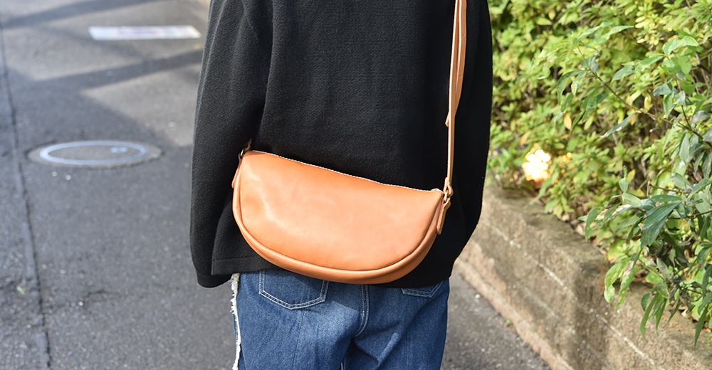 一款日本製HERZ職人手作皮革半圓馬鞍包，以褐色為主色調，由側面視角拍攝，包袋呈半月形，皮革質感明顯，搭配人物的黑色上衣和牛仔褲。