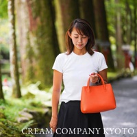 一位女性手持亮橙色手提托特包，包身為皮革材質，設計簡約。背景為自然景觀，有樹木與小徑。