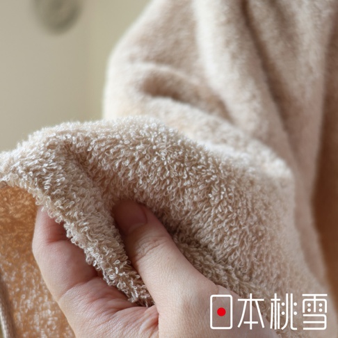 一隻手正在觸摸一條淺米色、柔軟的毛圈毛巾，展示其細緻的織物紋理。毛巾的材質看起來薄且蓬鬆。