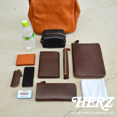 多款職人手作皮革配件，包含焦糖色皮革托特包、黑色化妝包、各式棕色系皮夾、筆袋、手機套及鑰匙包，搭配一瓶礦泉水及薄荷糖盒，呈現豐富的收納組合。