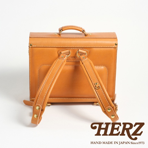 職人手作皮革橫式書包，淺棕色皮革，附有提把及兩條可調節肩帶，背面有壓印的HERZ品牌字樣及HAND MADE IN JAPAN Since1973字樣。