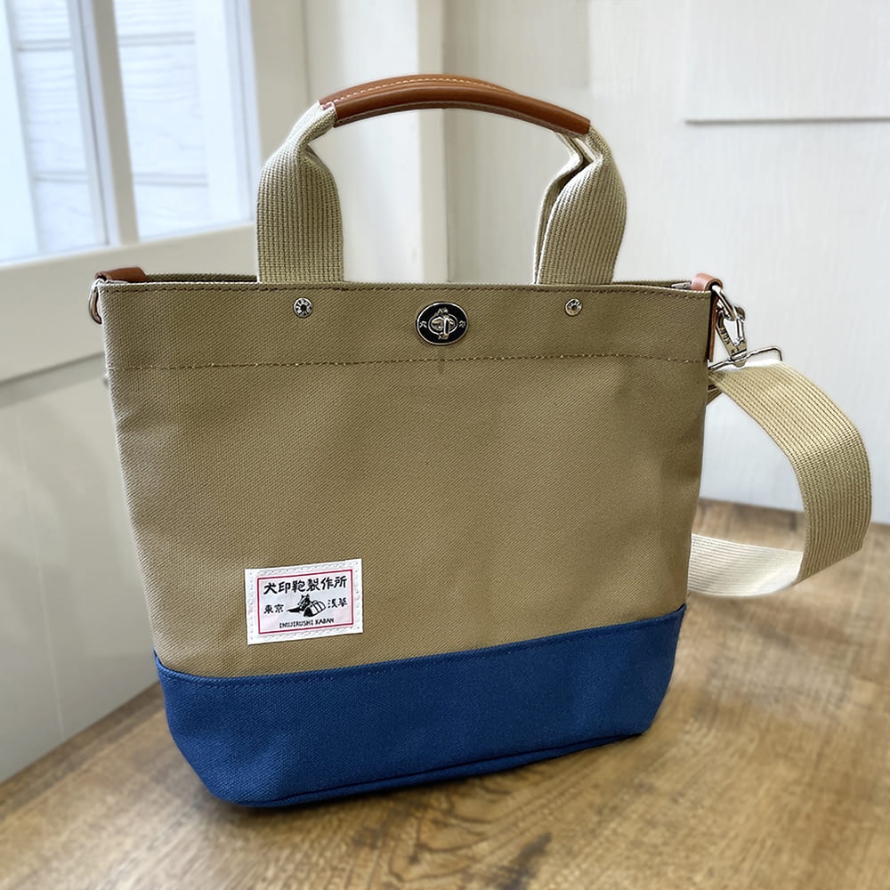 犬印純棉帆布兩用托特包，主體為卡其色，底部為海軍藍，配有咖啡色皮革提把與米色帆布可拆卸斜背帶，包身左前方有品牌標誌貼紙。