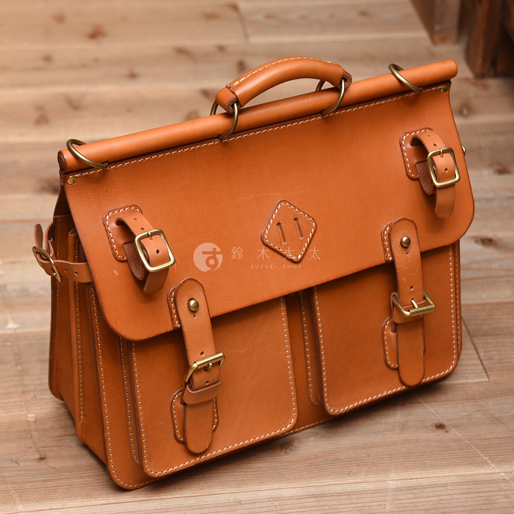 職人手作皮革旅行包 A-102，採用棕色皮革製成，設有頂部提把、可調節式皮帶和金屬扣環，以及前方雙口袋設計。