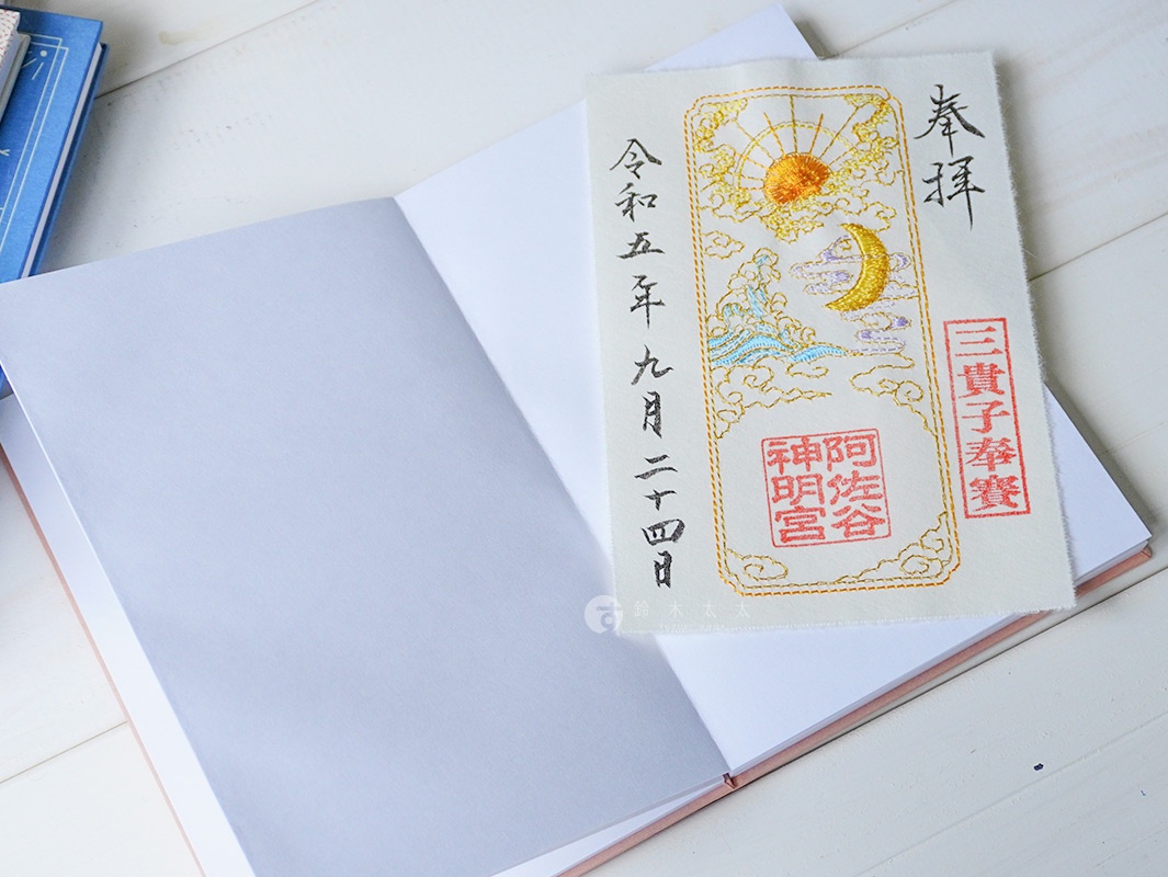 淺色和紙御朱印帳，攤開內頁。上方置放一塊白色布質御朱印，繡有金色太陽、彎月、祥雲、流水及日文文字、紅色印章。