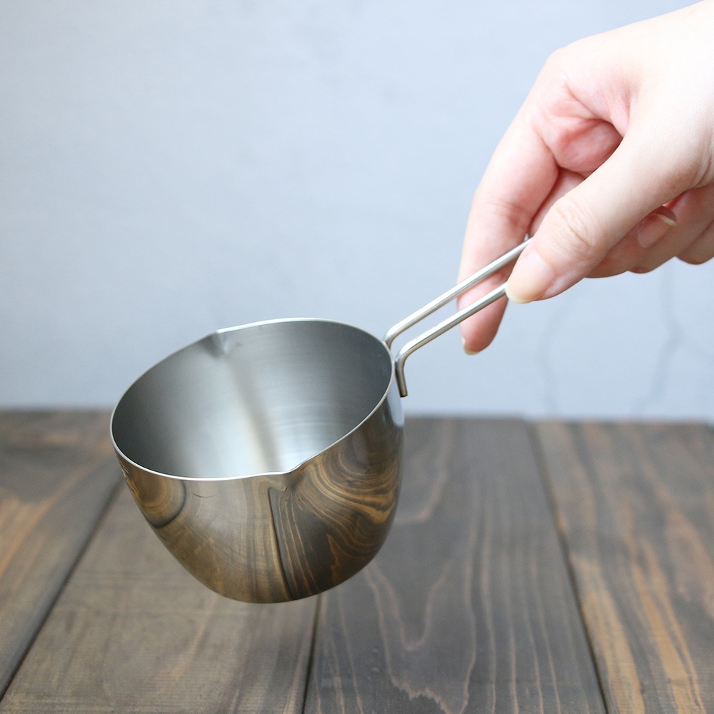 一只手正握著一個不鏽鋼長柄量杯，量杯為銀色，材質為不鏽鋼，帶有長柄，杯口有一處缺口設計，量杯放置在木紋桌面背景上。