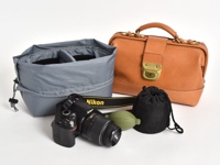 一個淺棕色皮革製的醫生包，配有金色鎖扣和提把。旁邊有一個灰色布製的相機收納袋，內有隔層。黑色單眼相機和綠色吹塵球放在前面。