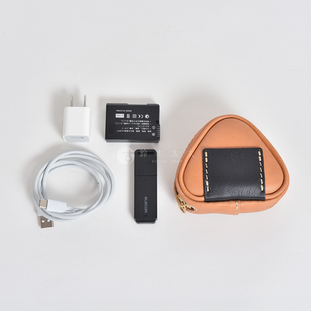 職人手作皮革三角飯糰造型小物收納包，淺棕色皮革，正面有黑色皮革裝飾與白色縫線，並設有金色拉鍊。旁邊擺放著白色充電頭、白色USB充電線、黑色電池與黑色USB隨身碟。