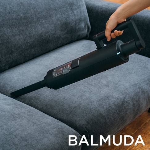 手持黑色 BALMUDA The Cleaner 無線吸塵器，吸嘴貼近灰色布沙發的縫隙，吸塵器本體為黑色，配有透明集塵筒和銀灰色按鈕。
