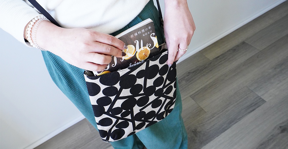 BAN INOUE 自然織紋麻布斜背包 (果實花紋)，斜背包內放有一本關於柑橘料理的書，一位女士手戴手鍊，身穿白色毛衣和綠色燈芯絨褲，正在調整書本的位置。