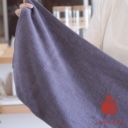一條深紫色、觸感柔軟的薄毛巾，表面有精緻的暗紋，材質為吸水性佳的棉質，由手部輕柔拿起，展現其輕盈質感。