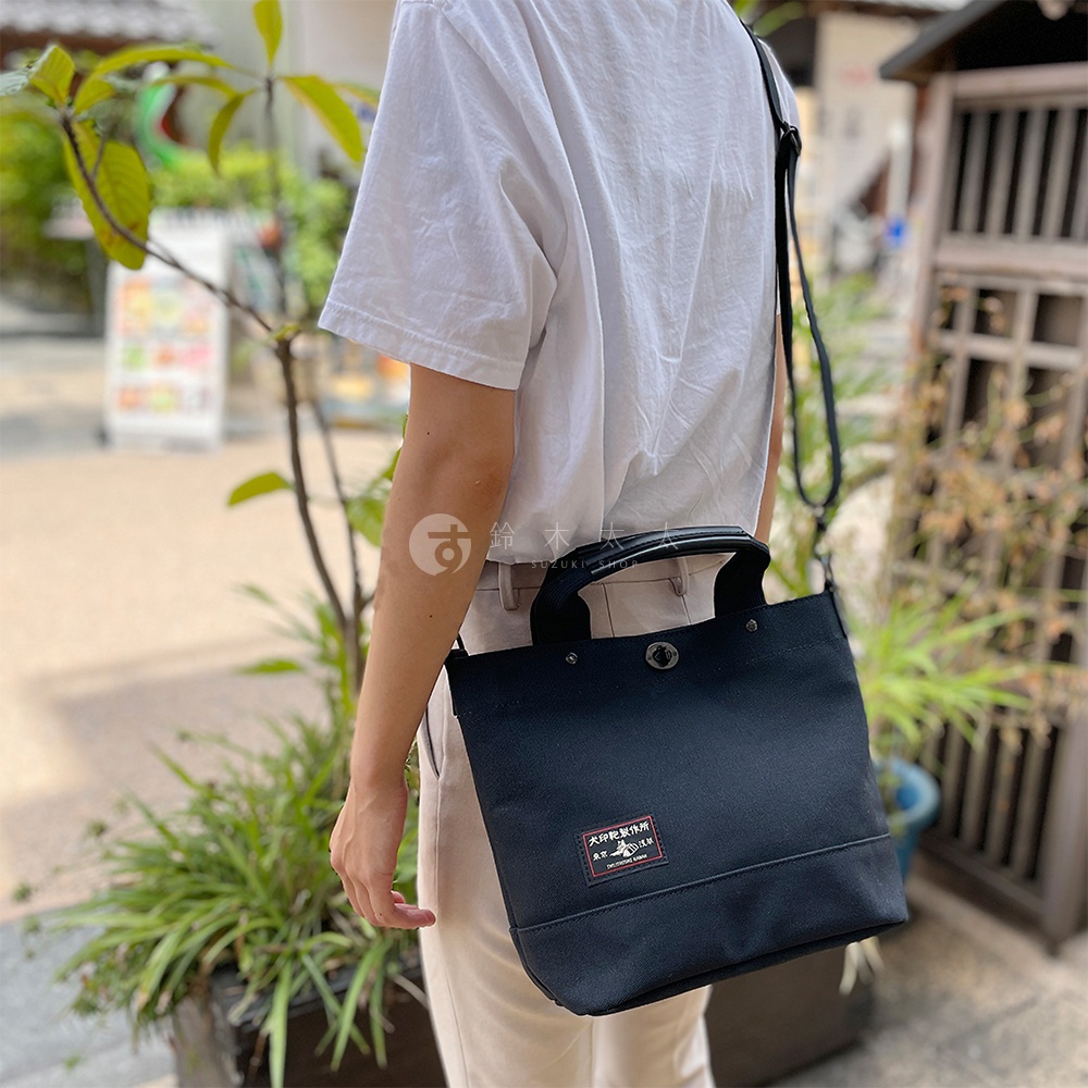 黑色帆布製成的兩用托特包，配有提把和可拆卸肩帶，包包正面有一個方形的紅色與白色標籤。