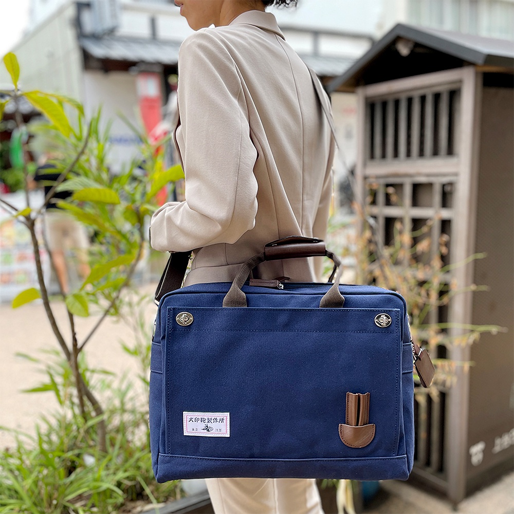 深藍色純棉帆布公事包，配有棕色皮革提把和裝飾，包包正面有品牌標籤及皮革配件，可肩背或手提。