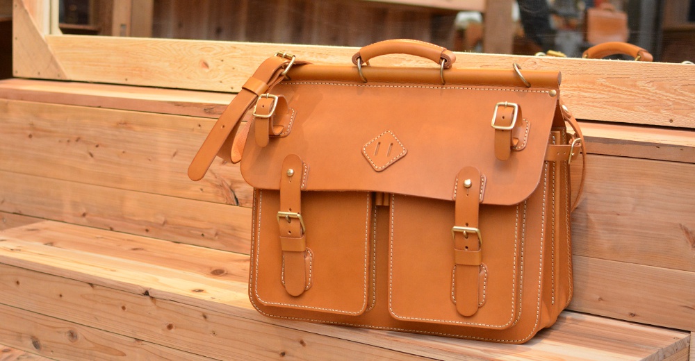職人手作皮革旅行包 A-102，採用鞣製皮革製成，主體為焦糖棕色，設有雙前袋、可調節肩帶和頂部提把，並以對比色縫線裝飾。