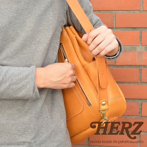 一名身穿灰色長袖上衣的人士背著日本職人品牌HERZ的淺褐色皮革斜背包，該包款呈現溫潤的皮革紋理，配有金屬拉鍊與厚實背帶，畫面右下方印有品牌名稱與日本手工製造標誌，背景為簡約的紅磚牆。