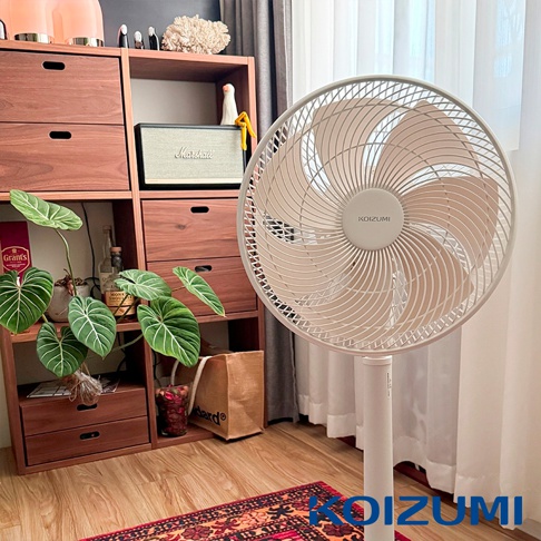 一台純淨白色的日本KOIZUMI品牌DC變頻遙控電風扇立於室內，風扇具有多葉片設計。背景為木質開放式收納櫃，櫃中擺放了Marshall音響、綠色盆栽與酒瓶等裝飾品，地板上鋪設印花地毯，整體呈現溫馨舒適的居家風格。