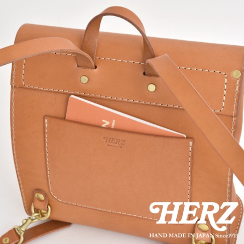 職人手作皮革翻蓋迷你後背包，採用淺棕色皮革，配有白色縫線及黃銅金屬扣件。包款正面有一個插袋，內有一本橘色筆記本，品牌標誌烙印在插袋上。