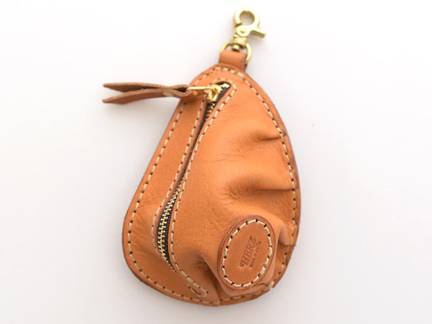 淺棕色皮革製成的餃子造型零錢包，帶有金色拉鍊和鑰匙圈扣環，縫線明顯，底部有圓形皮革標誌，標有「HERZ MADE IN JAPAN」。
