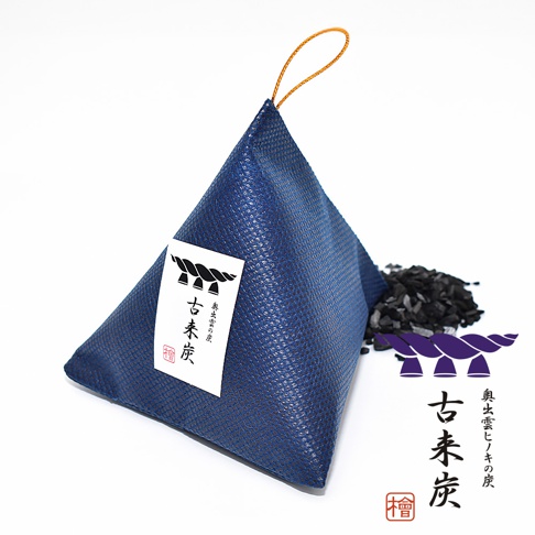 一個藍色三角錐狀的織物掛袋，表面帶有細緻紋理，頂端連接著金色的掛繩。袋子上貼有標籤，印有黑色注連繩圖案與古來炭字樣。袋子右側堆放著一些黑色木炭碎片，旁邊並排展示著相同的黑色注連繩圖案及古來炭品牌文字。