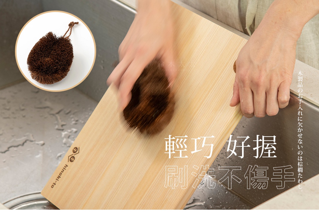 一隻圓形、深棕色的天然棕櫚毛刷，帶有打結的掛繩，正被一隻手握住，在一個淺色木製砧板上刷洗。