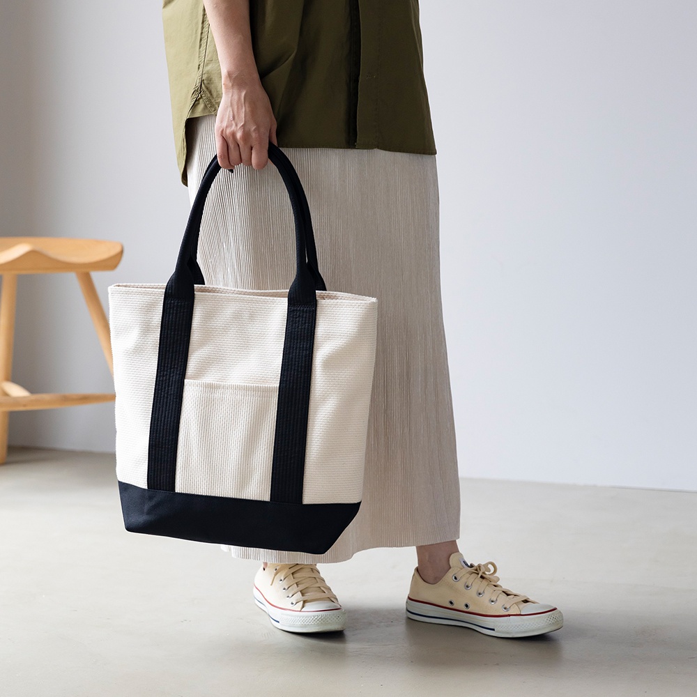 白色三河木綿刺子織托特包，搭配黑色織帶提把與底部，正面有一個黑色邊框的方形外袋。