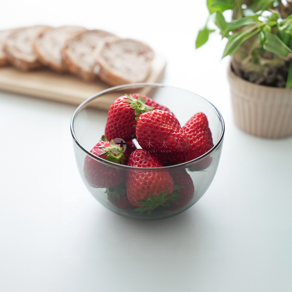 透明玻璃點心碗裝滿新鮮紅草莓，碗身圓潤，背景是切片的麵包和綠色植物。
