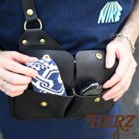 黑色皮革雙口袋斜背包，有兩個主要隔層，一個隔層內有藍白圖案的布料，另一個隔層內有黑色無線耳機。背包使用黃銅色金屬扣件。