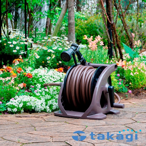 棕色捲盤上纏繞著棕色水管，水管末端連接黑色灑水槍，置於花園小徑上，周圍是盛開的各色花朵。