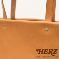 職人手作皮革托特包，呈現溫潤的淺棕色皮革紋理，包身有兩個金屬鉚釘點綴，提把為同色系皮革製作。