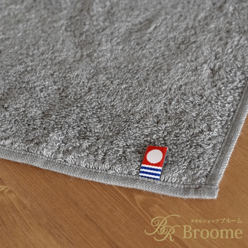 灰色長毛巾，材質柔軟，帶有今治品牌標誌，標誌為紅底白圓點，下方有藍色條紋。