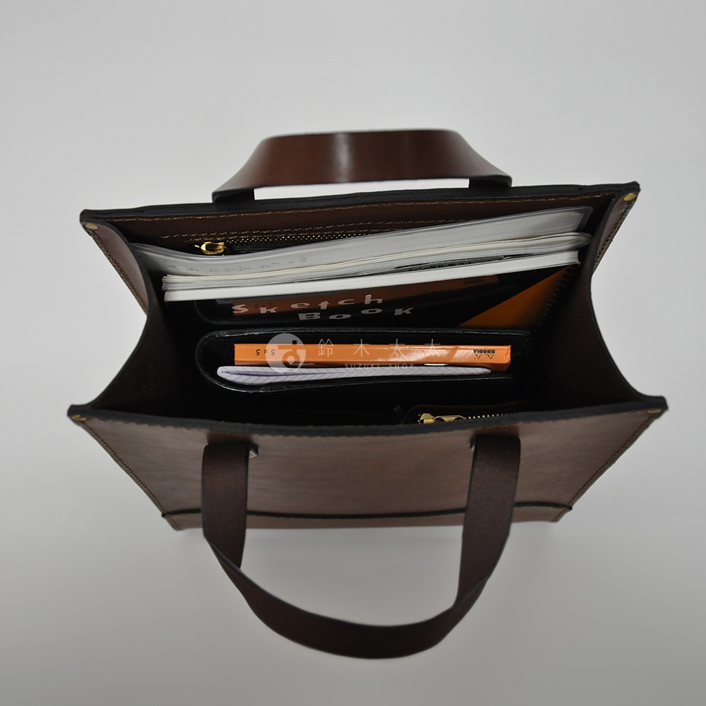 職人手作皮革直式托特包，棕色。包內裝有筆記本、皮夾和文件，整體呈現簡約實用的風格。