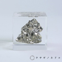 透明立方體中封存著一塊黃鐵礦，礦石呈現金屬光澤的銀灰色，表面有許多閃亮的結晶面，整體造型不規則，充滿立體感。