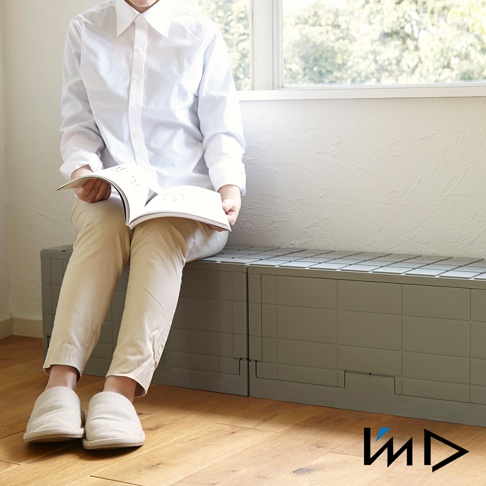 灰色的Grid格子磚可堆疊摺疊收納椅，由塑膠製成，具有方形紋理設計，可組合成長凳狀。一位穿著白色襯衫和米色褲子的人坐在一旁閱讀。