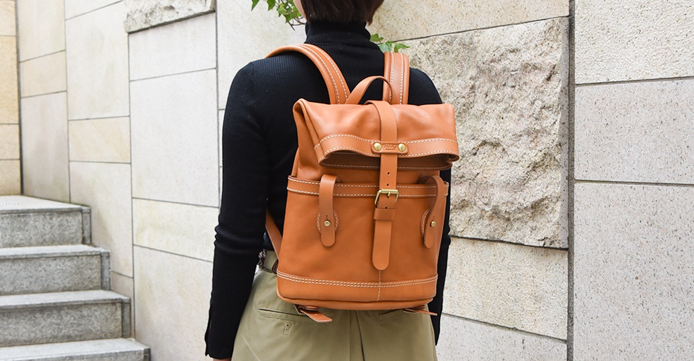 棕褐色皮革雙口袋折蓋後背包，配有可調節肩帶、皮帶扣開合及側邊皮帶細節，展現職人手作質感。